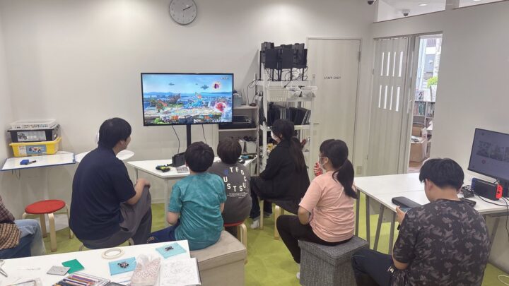 子どもがスタッフや他の子どもと一緒に活動しているシーン（プログラミングやゲームなど）。和やかで安心感のある雰囲気。顔は写らないか後ろ姿推奨
