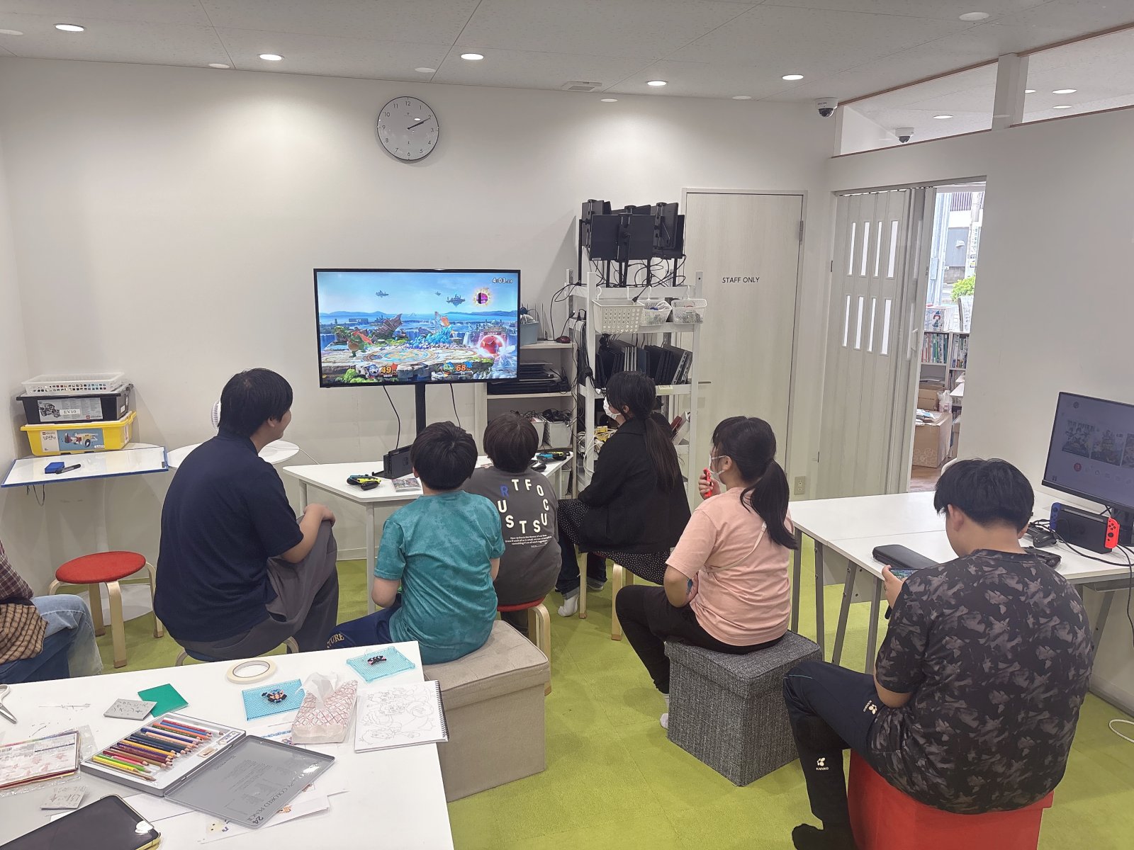 子どもがスタッフや他の子どもと一緒に活動しているシーン(プログラミングやゲームなど)。和やかで安心感のある雰囲気。顔は写らないか後ろ姿推奨