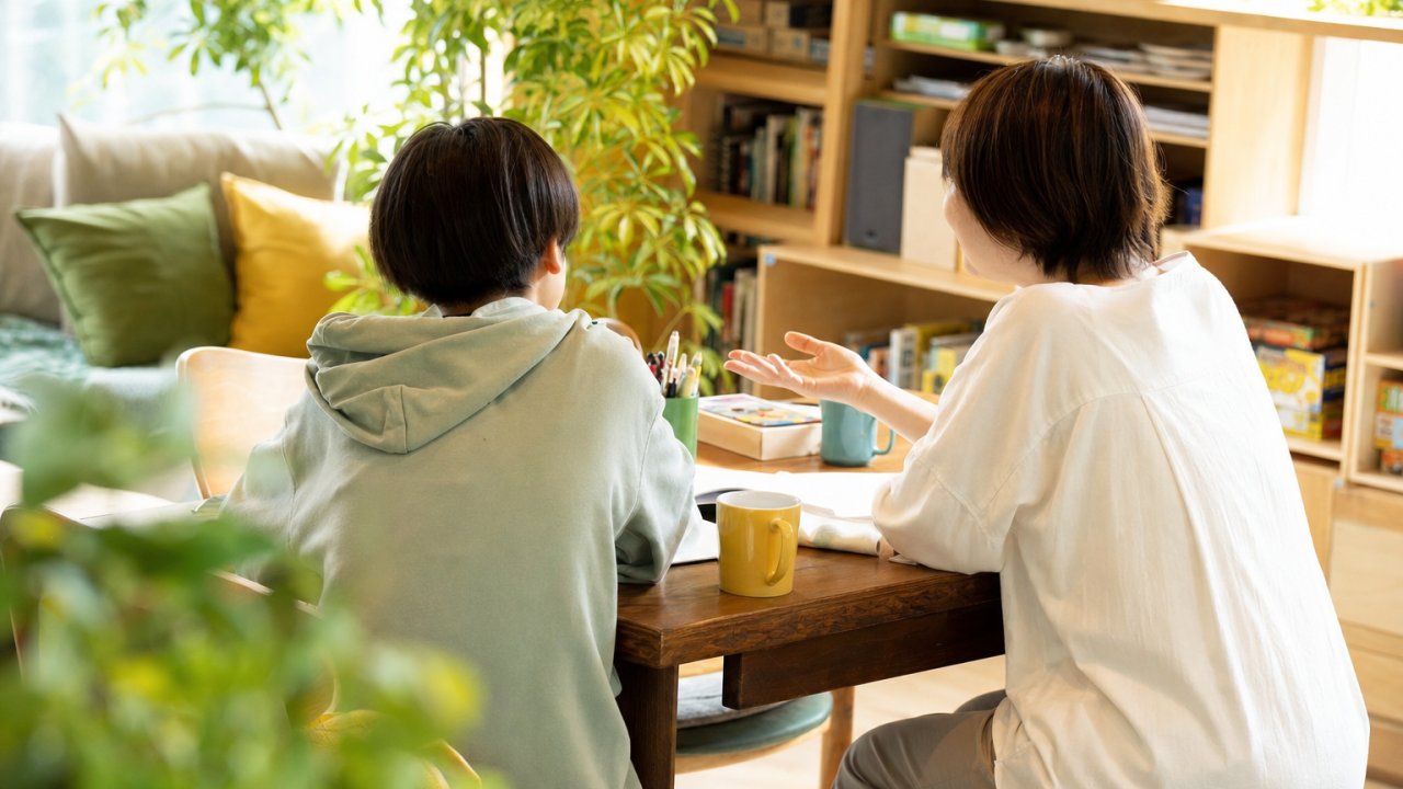 子どもが大人と横並びに座って、穏やかな表情で話しているシーン。カフェや家のリビングのような、くつろいだ空間のイメージ