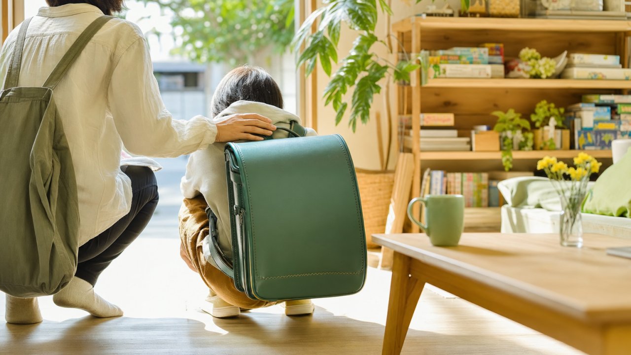 朝の玄関先で、ランドセルを背負ったまましゃがんでいる子どもと、そっと横に寄り添う大人（顔は映らなくてOK）。ランドセルの色は何色でもOK